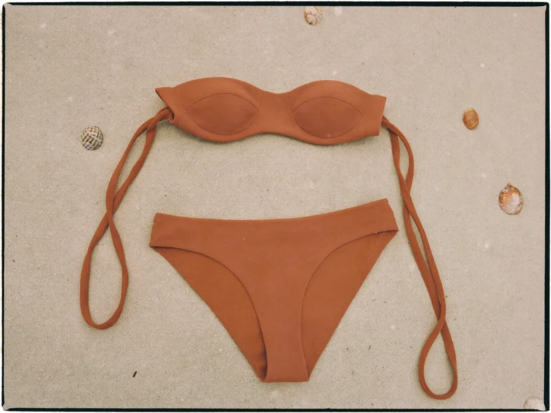Bikini terracotta in tinta unita adagiato sulla sabbia con piccole conchiglie
